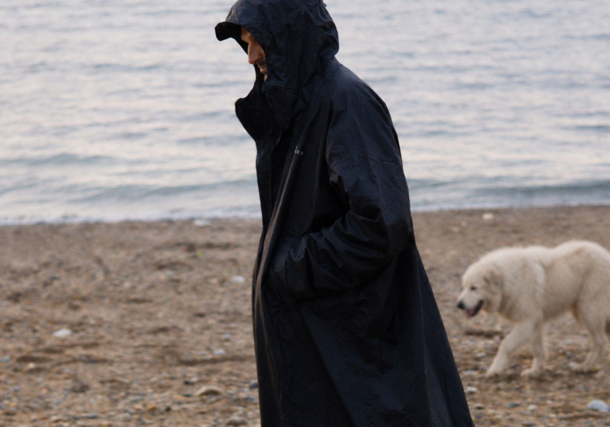 A person wearing a long black VOITED Allweather Coat stands on a pebble beach with the ocean in the background, and a white dog walks nearby. AI modified