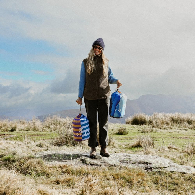Smiling woman on a hilltop holding two packed VOITED Cloudtouch blankets in their stuff sacks.