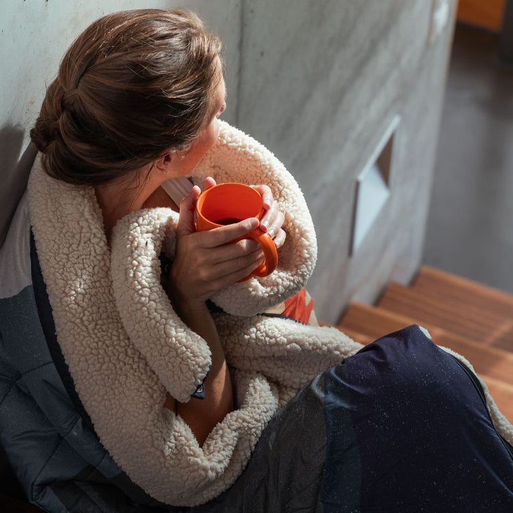 A woman sits on a wooden bench wrapped in a cream sherpa fleece Cloudtouch blanket, holding an orange mug. She is wearing a gray quilted puffer jacket and a white top, set against a concrete wall and wooden stairs.VOITED