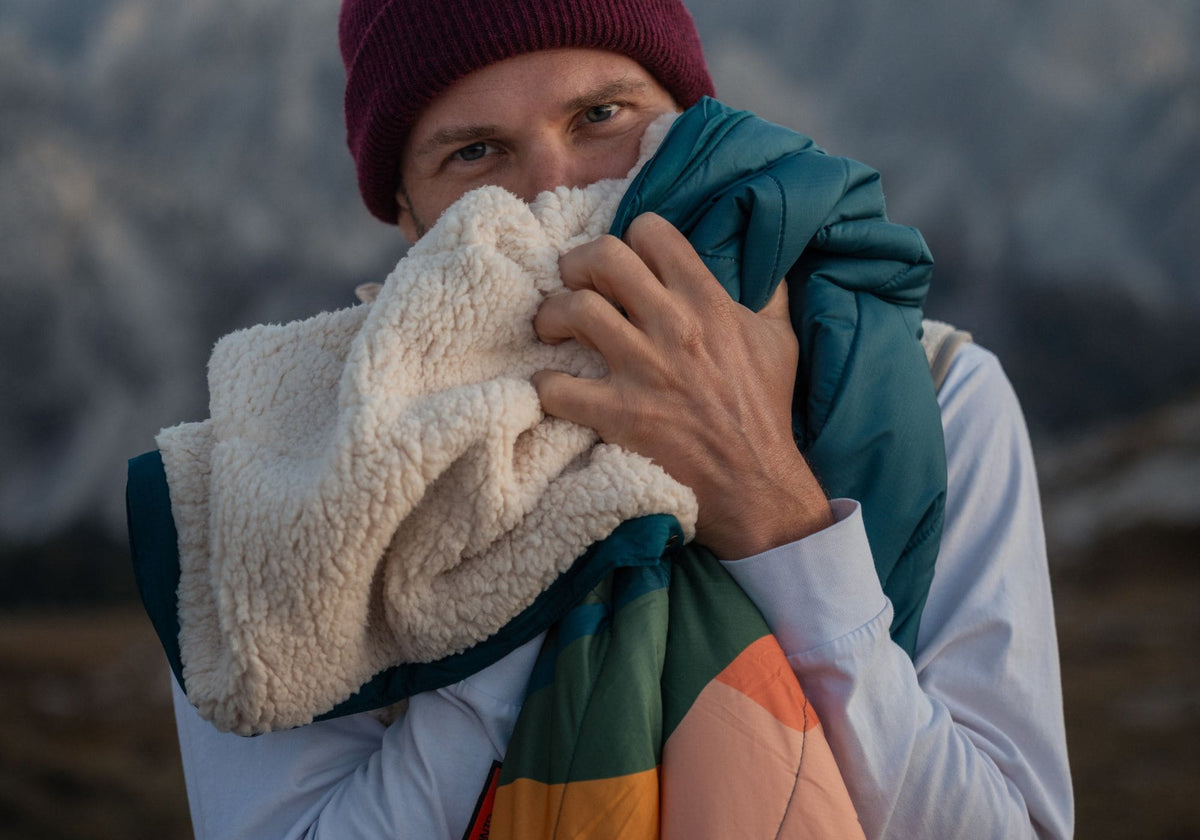 A man outdoors in a mountain setting, wearing a burgundy beanie, holds up a VOITED Cloudtouch Blanket with a cream sherpa fleece side and a teal and orange side, with mountains blurred in the background."